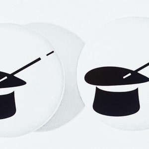 May include: Two white round coasters with a black and white illustration of a top hat and a magic wand. The top hat is sitting on top of a table and the magic wand is sticking out of the top hat.