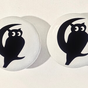May include: Two white round buttons with black owl silhouettes perched on a crescent moon. The owls have large, round eyes.