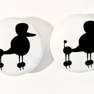 May include: Two white round ceramic buttons with black silhouette images of poodle dogs. The poodles are facing right and have a curly tail.