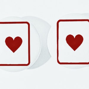 May include: Two white coasters with a red heart design inside a red square. The coasters are round and have a slightly raised edge.
