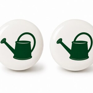 May include: Two round white ceramic cabinet knobs, each featuring a dark green graphic illustration of a watering can centered on the front surface. The knobs have a smooth, glossy finish and a simple, functional design for furniture hardware.