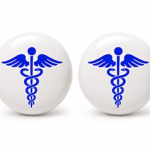 May include: Two round white ceramic cabinet knobs, each featuring a centered blue caduceus symbol. The medical emblem consists of a winged staff with two snakes intertwined around it, printed in a bold, solid blue color on the smooth surface.