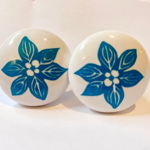 May include: Two white ceramic knobs with a blue floral design. The flowers have five petals and a white center with three white dots.