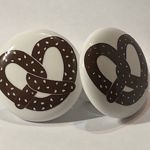 May include: Two white ceramic knobs with a brown pretzel design. The pretzels have a dotted pattern.