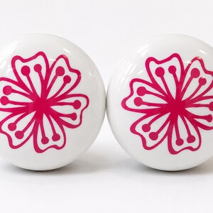 May include: Two round, white ceramic cabinet knobs, each featuring a vibrant, hot pink floral graphic printed on the front. The flower design consists of six symmetrical petals with rounded tips and small circular accents at the end of each petal.