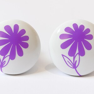 May include: Two white ceramic cabinet knobs with a glossy finish. Each knob features a purple daisy design with petals and leaves. The knobs are round and suitable for drawers or cabinets.