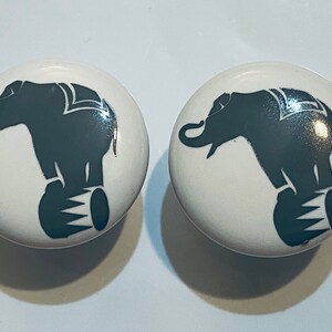 May include: Two white ceramic knobs with a black silhouette of an elephant balancing on a ball. The elephant has a trunk raised in the air.