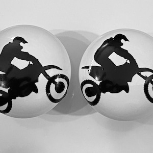 May include: Two white ceramic knobs with a black silhouette of a person riding a dirt bike.