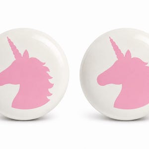 May include: Two round white cabinet knobs, each featuring a centered pink silhouette of a unicorn head in profile. The unicorn design includes a pointed horn, a mane, and an ear, rendered in a solid matte pink color against the smooth white surface.