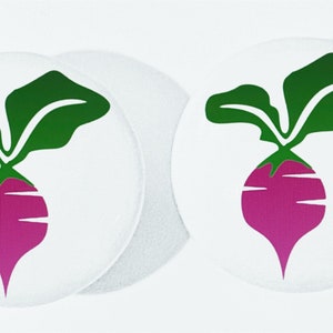 May include: Two white round buttons with a graphic of a purple beet with green leaves. The beet has a long, tapered shape and is centered on the button. The leaves are a vibrant green, and the beet is a deep purple.