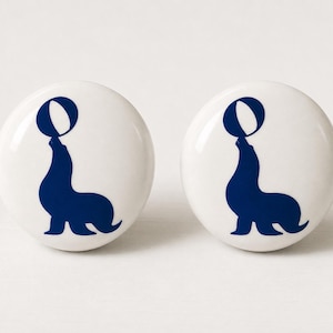 May include: Two round white ceramic cabinet knobs, each featuring a dark blue silhouette graphic of a sea lion balancing a ball on its nose. The knobs have a smooth, glossy finish and a circular shape, providing a clean and simple design element.