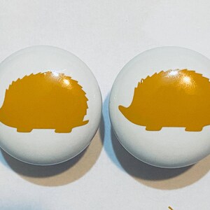 May include: Two white ceramic knobs with a yellow hedgehog silhouette design.
