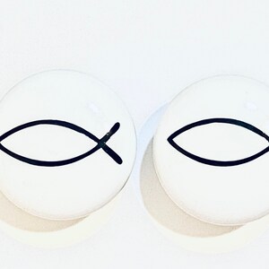 May include: Two white ceramic round ornaments with a black outline of the Christian ichthys symbol, also known as the Jesus fish.