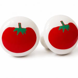May include: Two round white ceramic cabinet knobs, each featuring a vibrant red tomato illustration with a green stem and leaves centered on the front surface. The knobs have a smooth, glossy finish and a spherical shape, designed for furniture hardware.