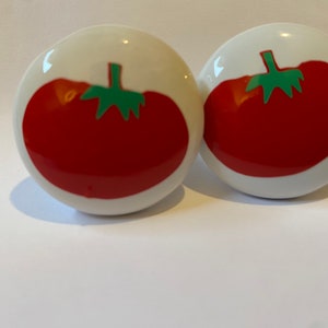 May include: Two white ceramic knobs with red tomato designs. The tomatoes have green stems and leaves.