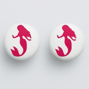 May include: Two white, round cabinet knobs with a magenta mermaid silhouette design. The mermaid has flowing hair and a fishtail. The knobs are suitable for drawers or cabinets.