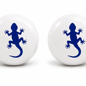 May include: Two spherical white knobs, each featuring a centered dark blue silhouette of a lizard. The smooth, round objects have a glossy finish and display a clear, high-contrast graphic of the reptile on their front-facing surface.