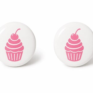 May include: Two circular white pin-back buttons, each featuring a centered graphic of a pink cupcake topped with a single cherry. The design is simple and clean, with the pink cupcake illustration standing out against the solid white background.