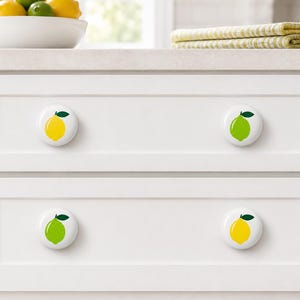 May include: Four round white cabinet knobs installed on two white drawers. Each knob features a graphic illustration of a single citrus fruit with a green leaf. Two knobs display a yellow lemon, and two knobs display a bright green lime.