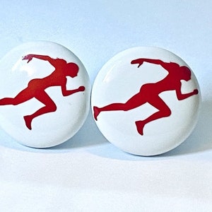 May include: Two white cabinet knobs with a red silhouette of a runner in motion. The knobs are round and have a glossy finish, suitable for a sports-themed decor.