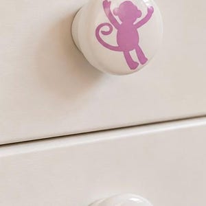 May include: Two round white ceramic drawer knobs, each featuring a smooth glossy finish and a centered light purple silhouette graphic of a monkey with its arms raised and a curled tail, attached to a light cream-colored wooden furniture surface.
