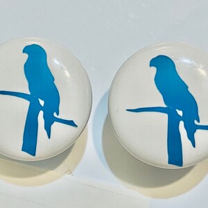 May include: Two white cabinet knobs with a glossy finish. Each knob features a blue silhouette of a bird perched on a branch. The bird and branch are a solid, vibrant blue against the white background. The knobs are round and appear to be made of ceramic.