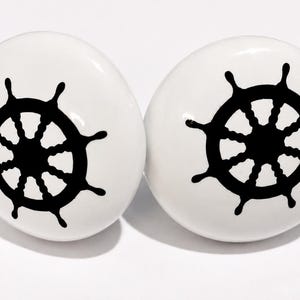 May include: Two round, white ceramic cabinet knobs, each featuring a bold, black graphic of a ship's steering wheel centered on the surface. The knobs have a smooth, glossy finish and a domed shape, providing a clean, high-contrast nautical design.