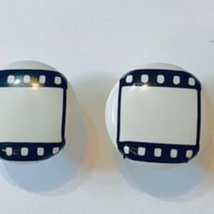 May include: Two white ceramic knobs with a black film strip design. The knobs are round and have a smooth surface.