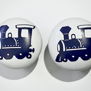 May include: Two white ceramic cabinet knobs with a navy blue train design. The train illustration features a locomotive with a chimney, windows, and wheels. These knobs are suitable for drawers or cabinets.