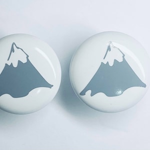 May include: Two white ceramic knobs with a gray mountain design. The knobs are round and have a glossy finish.