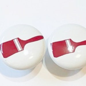 May include: Two white ceramic knobs with red paintbrush designs. The paintbrushes are outlined in white.