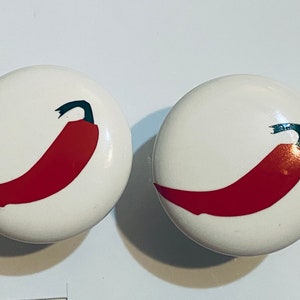 May include: Two white ceramic knobs with red chili pepper designs. The chili peppers are outlined in black and have a green stem.