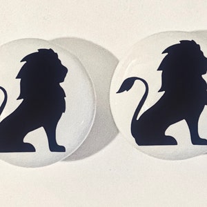 May include: Two white ceramic round buttons with a black silhouette of a lion on each. The lion is facing to the right and has a mane.