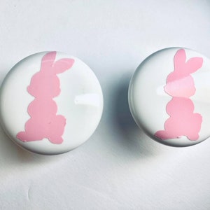 May include: Two white ceramic knobs with pink bunny silhouettes. The knobs are round and have a smooth surface.