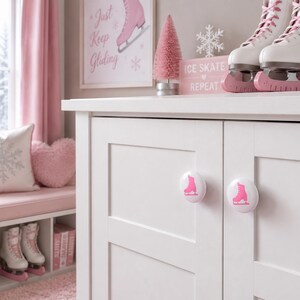 May include: A white cabinet featuring two round knobs, each decorated with a pink ice skate graphic. The cabinet doors are flat-paneled, and the knobs are positioned centrally on each door, providing a functional and decorative accent to the furniture.