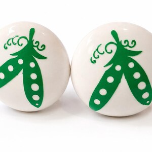 May include: Two round, white ceramic cabinet knobs, each featuring a vibrant green graphic of a pea pod with visible circular peas inside and small curly tendrils at the top. The surface of the knobs is smooth, glossy, and spherical in shape.