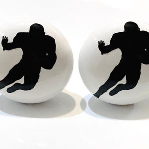May include: Two white ceramic knobs with black silhouettes of football players in a running pose.