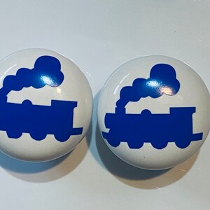 May include: Two white ceramic knobs with a blue train silhouette design. The train has a blue smoke plume and is facing right.