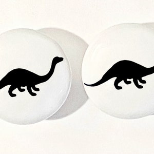 May include: Two white round buttons with a black silhouette of a dinosaur on each. The dinosaur is facing right and has a long neck.