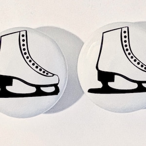 May include: Two white round buttons with black outlines of ice skates. The skates have a white boot with black laces and a black blade.
