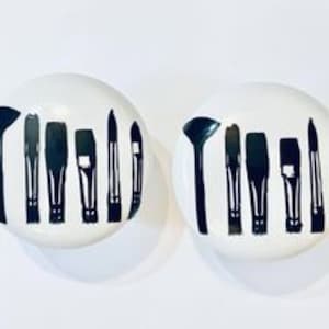 May include: Two white round ceramic buttons with black paint designs of paint brushes. The buttons are about 1 inch in diameter.