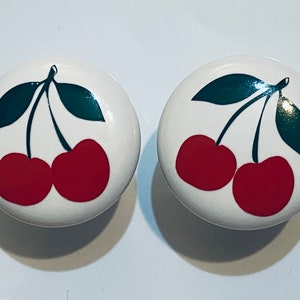 May include: Two white ceramic knobs with a red cherry design. Each knob features two cherries with green leaves.
