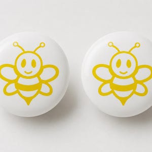 May include: Two circular white buttons, each featuring a centered yellow graphic illustration of a cartoon bee with two antennae, two wings on each side, and a striped body. The design is simple, clean, and symmetrical, presented on a plain white background.