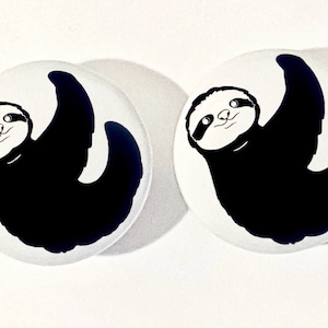 May include: Two white round buttons with black and white images of sloths hanging upside down. The sloths have black bodies and white faces with black eyes and mouths.