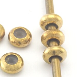 Pode incluir: Cinco contas de metal douradas com centros de vidro transparente. As contas são enfiadas em uma haste de metal fina.