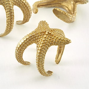 May include: Three gold-colored starfish-shaped napkin rings. The rings are made of metal and have a textured surface.