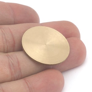 Op de afbeelding: Een klein, goudkleurig, cirkelvormig metalen object met een geribbelde rand en een getextureerd oppervlak. Het object wordt in de hand van een persoon gehouden tegen een witte achtergrond. Het lijkt een kleine schijf of component te zijn.