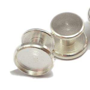 May include: Three silver-toned metal settings for a jewelry making project. Each setting has a round, flat top with a raised edge and a small, round post in the center.