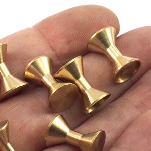 Puede incluir: Seis bobinas de atado de moscas de metal de color dorado con parte superior e inferior acampanada. Las bobinas están en una mano.