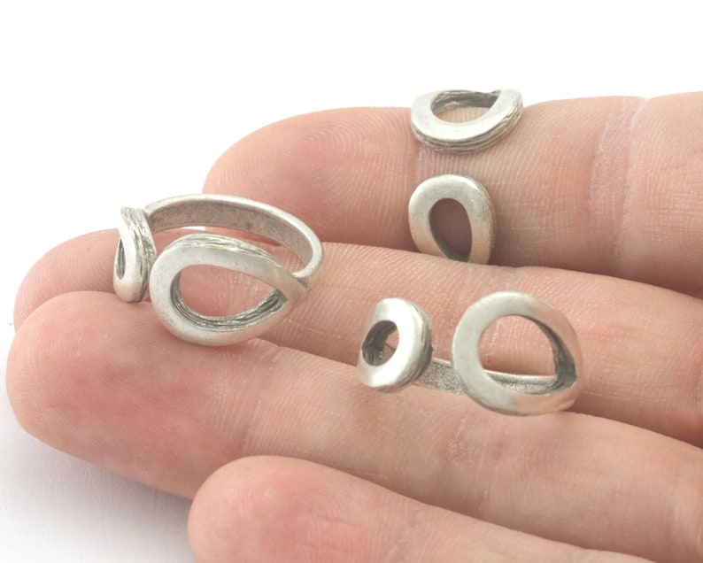 May include: Several silver-colored rings with an open teardrop design are displayed. The rings have a textured, matte finish. The rings are on a person's hand.